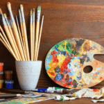 A paint pallet with a multitude of colors on it. To the left is a white container with about 15 wood handle artist paint brushes.